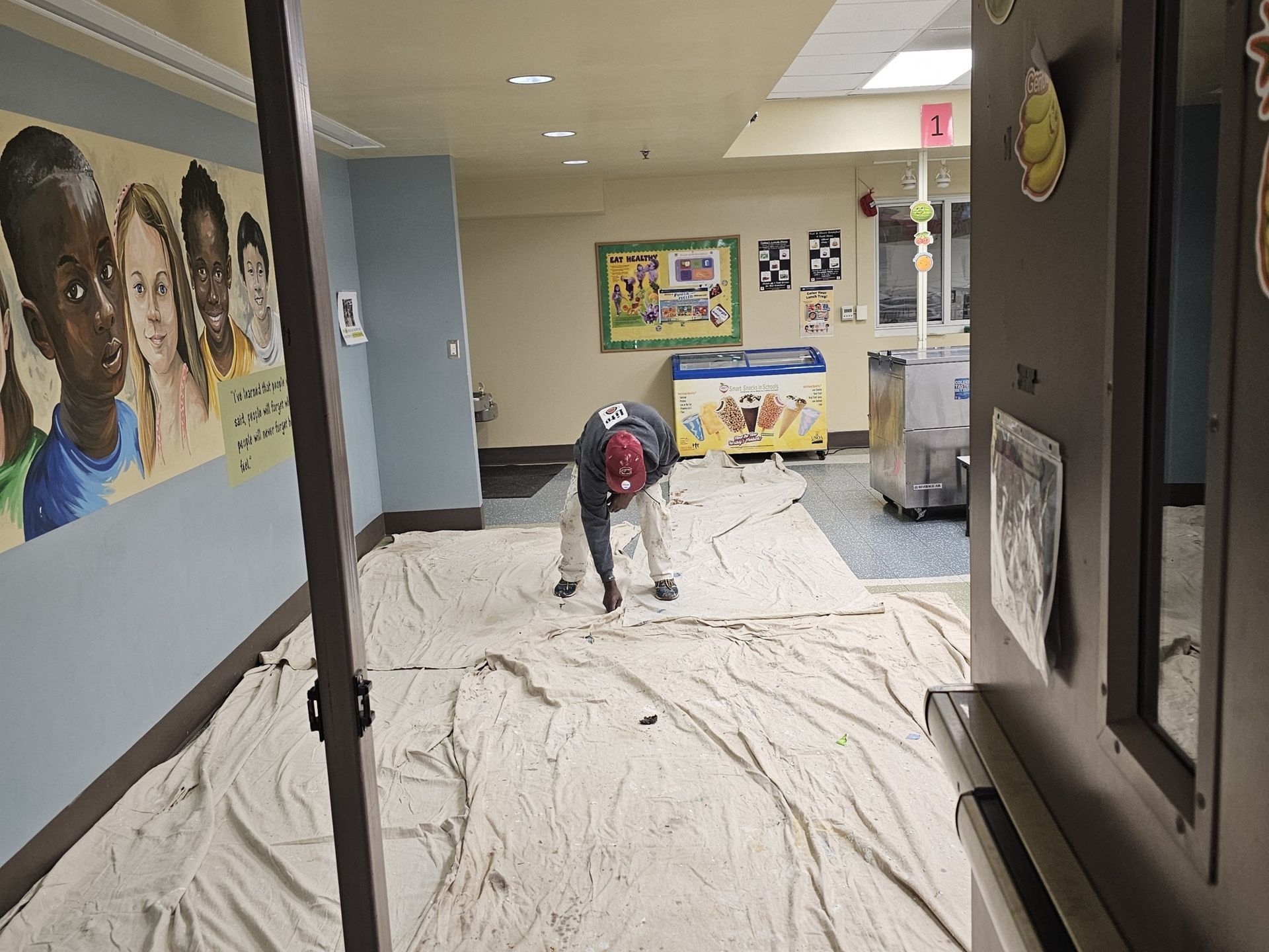 A person painting a hallway, standing on drop cloths. There is artwork and a doorway visible.