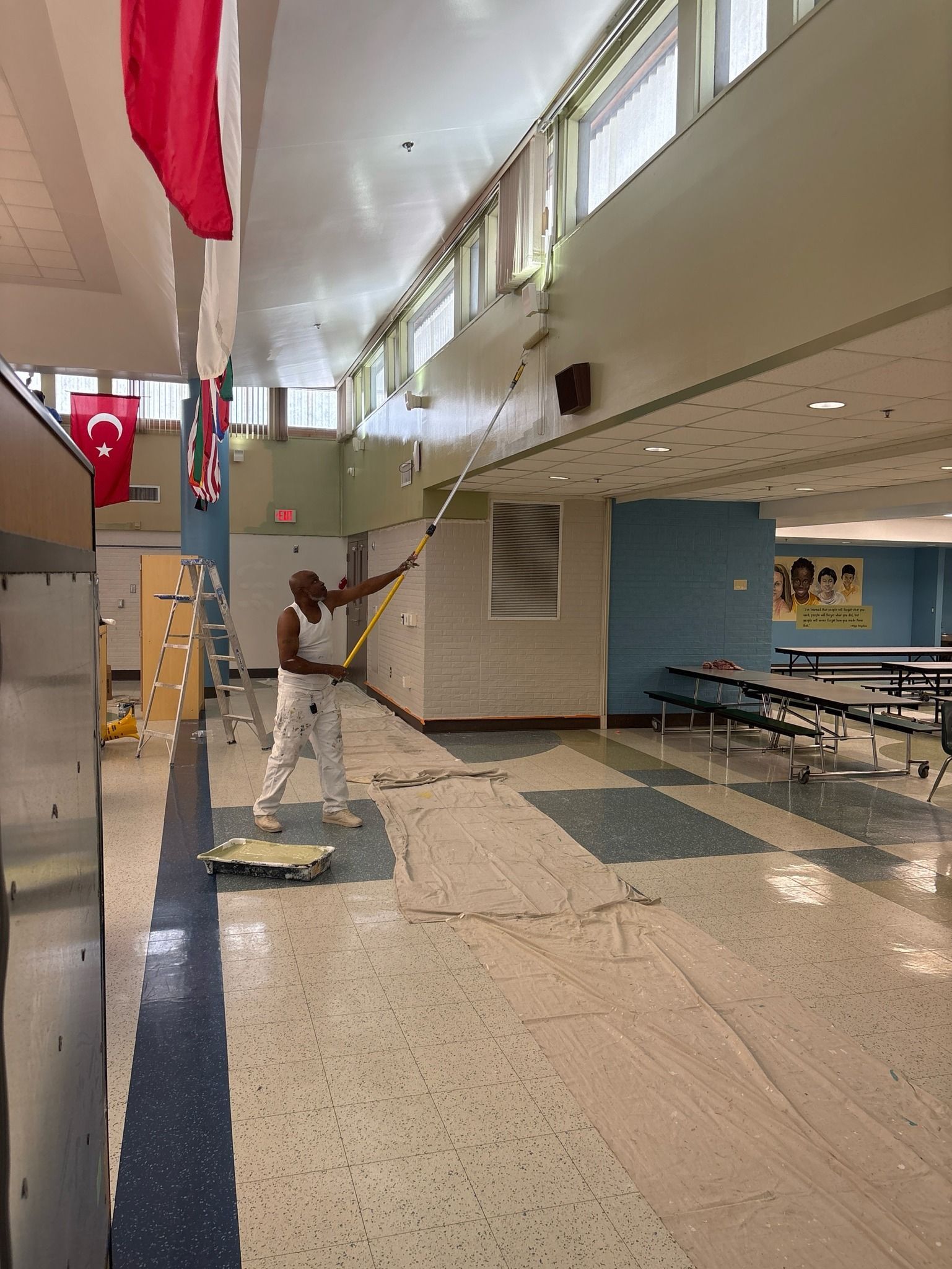 Man in a school hallway painting.  White paint covers the floor. Flags hang, lunch tables visible.