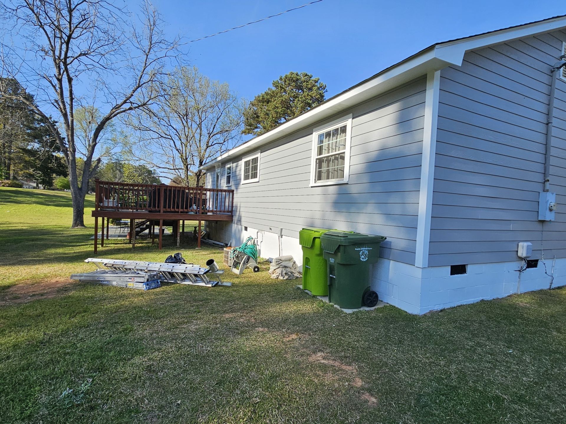 A gray house with a wooden deck in a grassy yard, with green trash cans nearby.