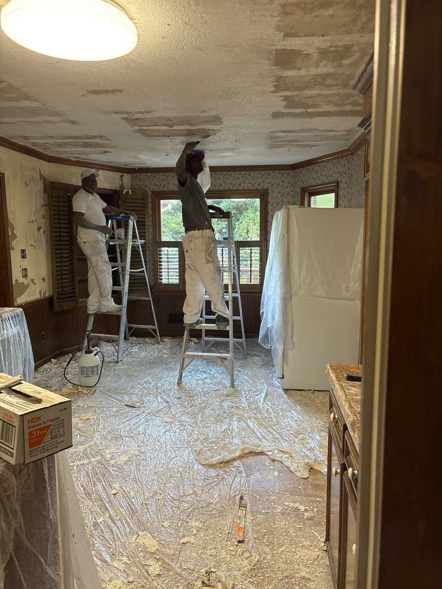 Three people on ladders scrape ceiling, flakes on floor. Kitchen renovation.