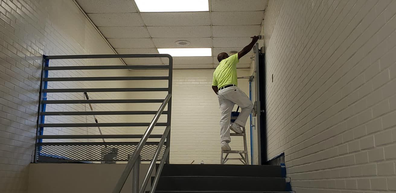 A painter on a ladder painting a white wall next to a staircase with a metal railing.