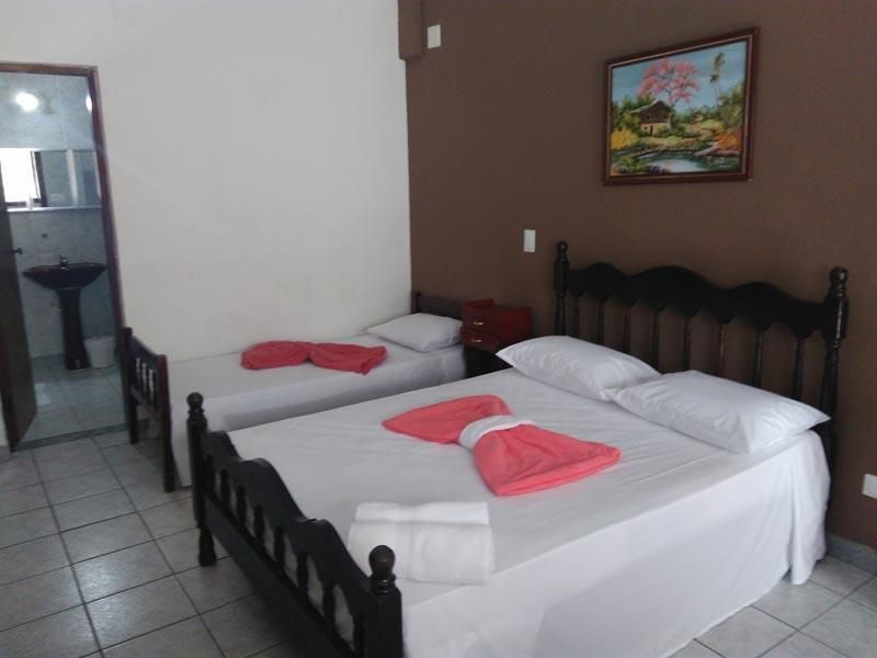Um quarto com uma cama de casal e uma cama de solteiro, ambas com lençóis brancos, detalhes em rosa e cabeceiras de madeira preta.
