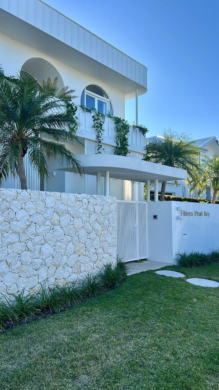 A modern, two-story white building with an arched design, stone wall, and palm trees under a clear blue sky.