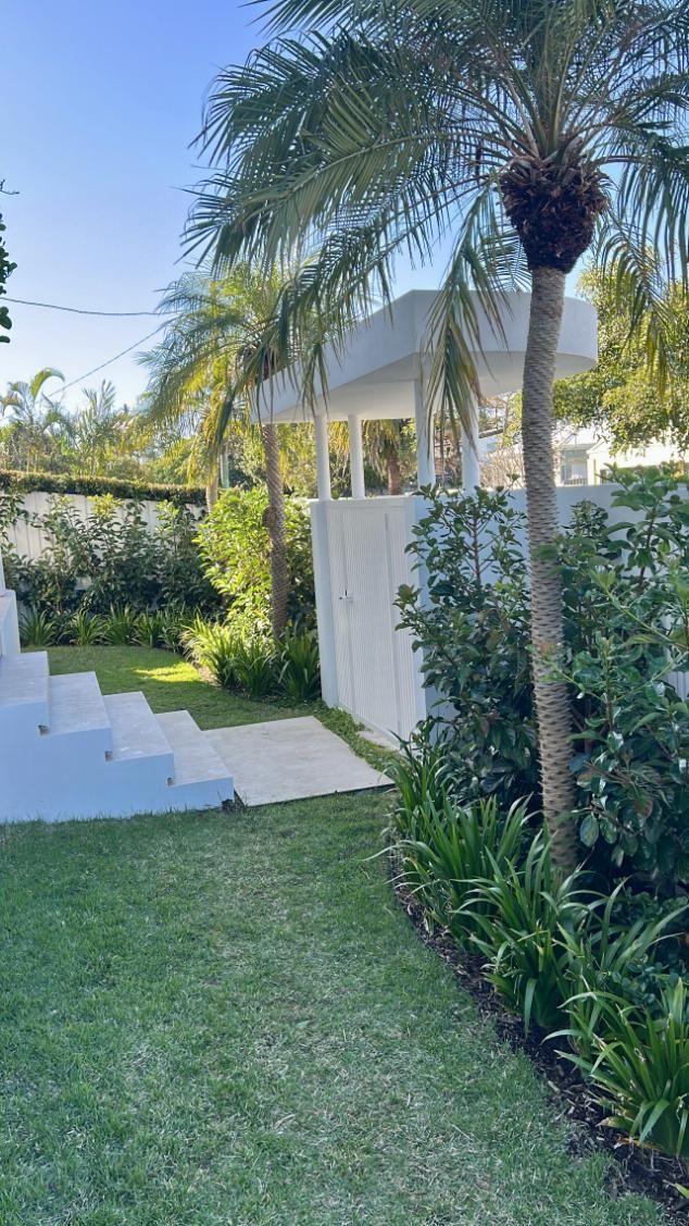 A grassy lawn leading to a set of white outdoor stairs next to a small white structure surrounded by trees and plants.