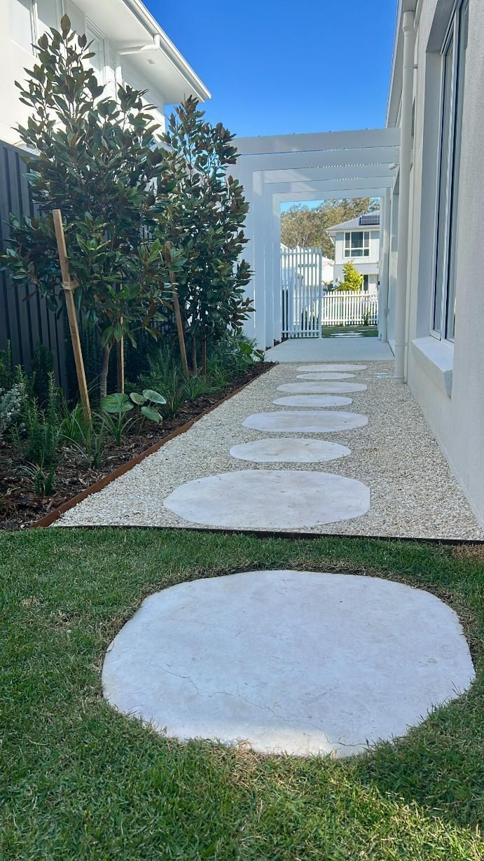 Large circular concrete stepping stones lead through a gravel garden path next to a white house under a blue sky.