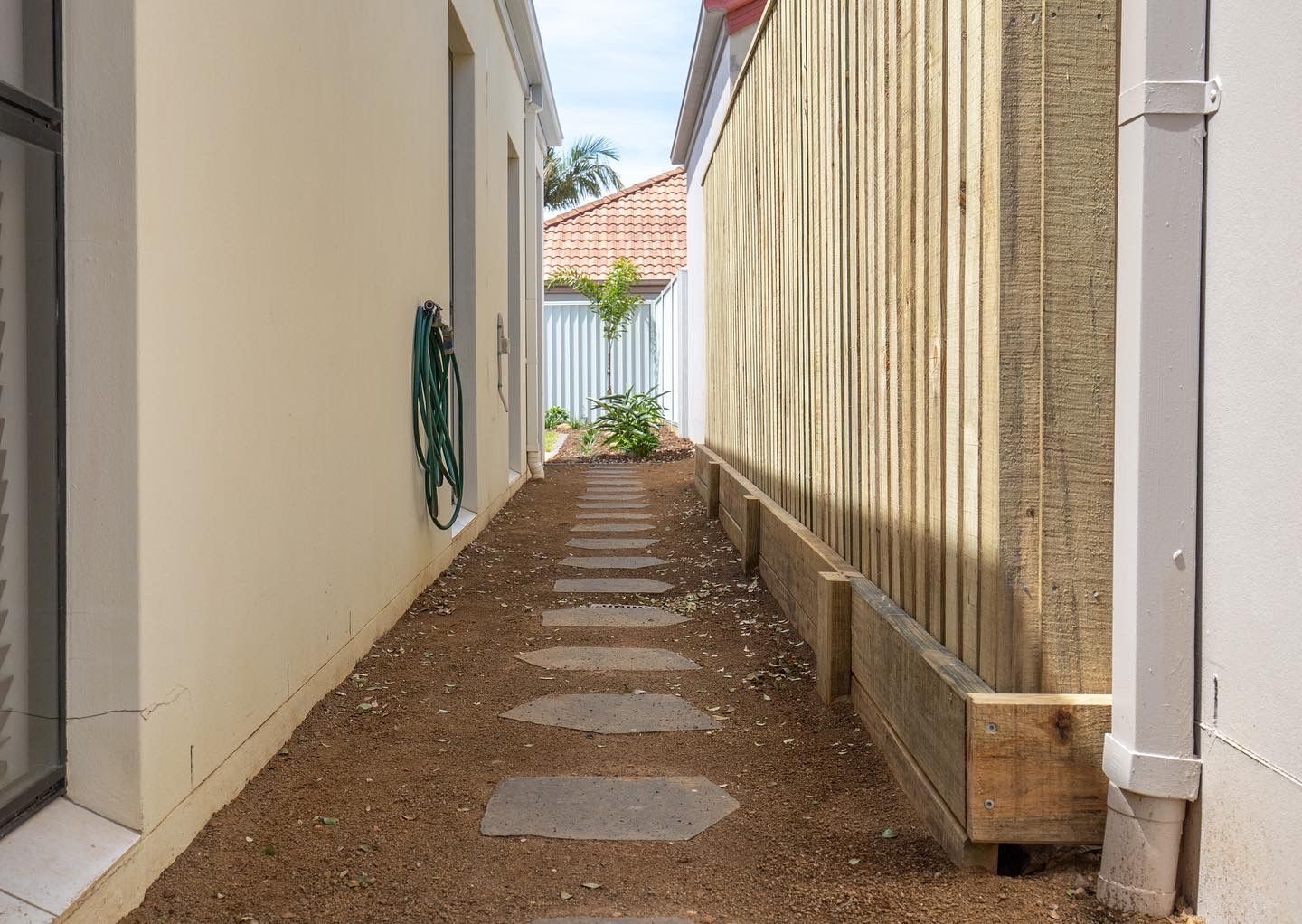 A narrow side yard with a path of stone pavers set into mulch, bordered by a wooden fence and a light-colored house wall.