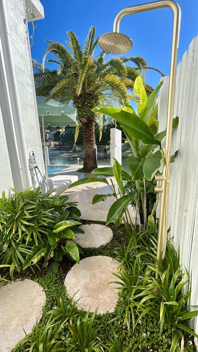 An outdoor gold shower stands in a tropical garden with stone stepping stones leading to a pool under a bright blue sky.