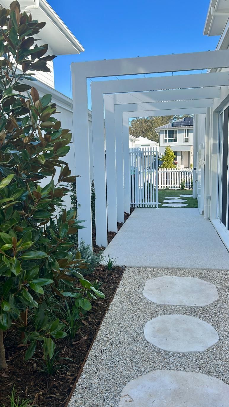 A white pergola spans a gravel path with round stepping stones, leading toward a house and white picket fence.