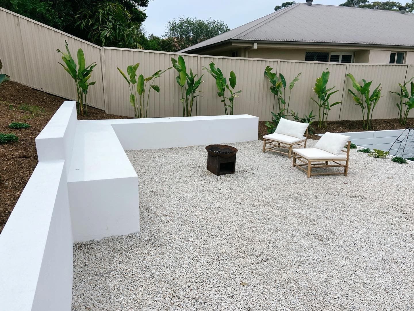 A modern outdoor seating area with a white L-shaped concrete bench, two lounge chairs, and a fire pit on gravel.
