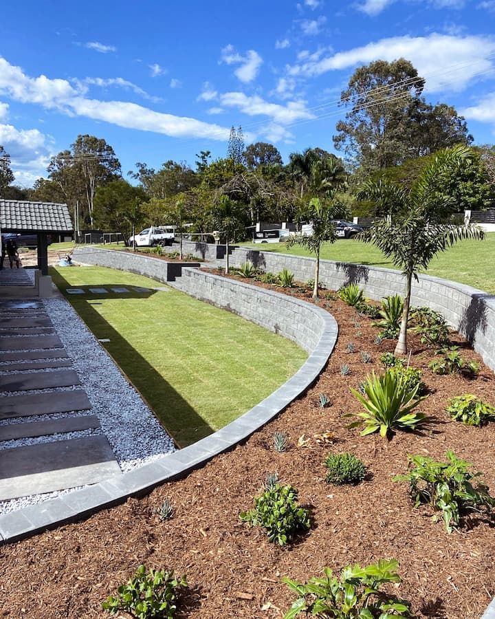 A landscaped backyard featuring tiered grey stone retaining walls, a curved lawn, mulch garden beds, and stone pathing.