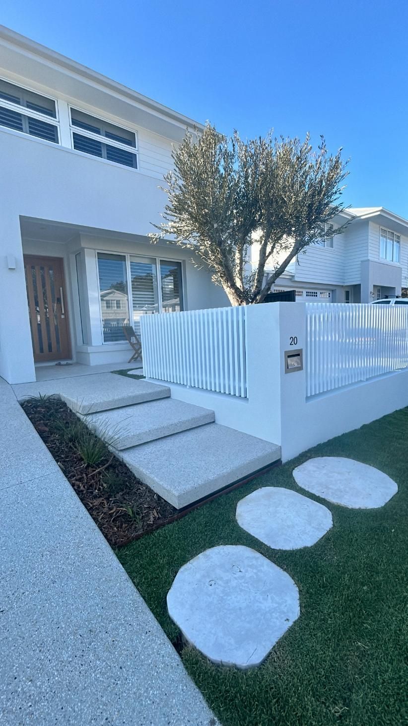 Modern white two-story house with a wooden front door, tiered concrete steps, white fencing, and round garden pavers.