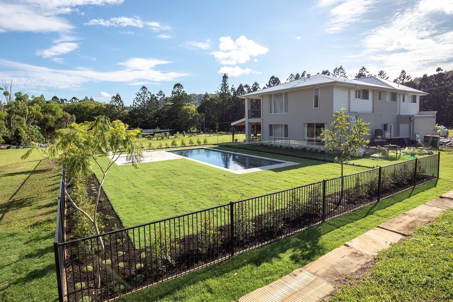 A two-story light gray house with a backyard pool, green lawn, and dark metal fence under a blue sky with light clouds.