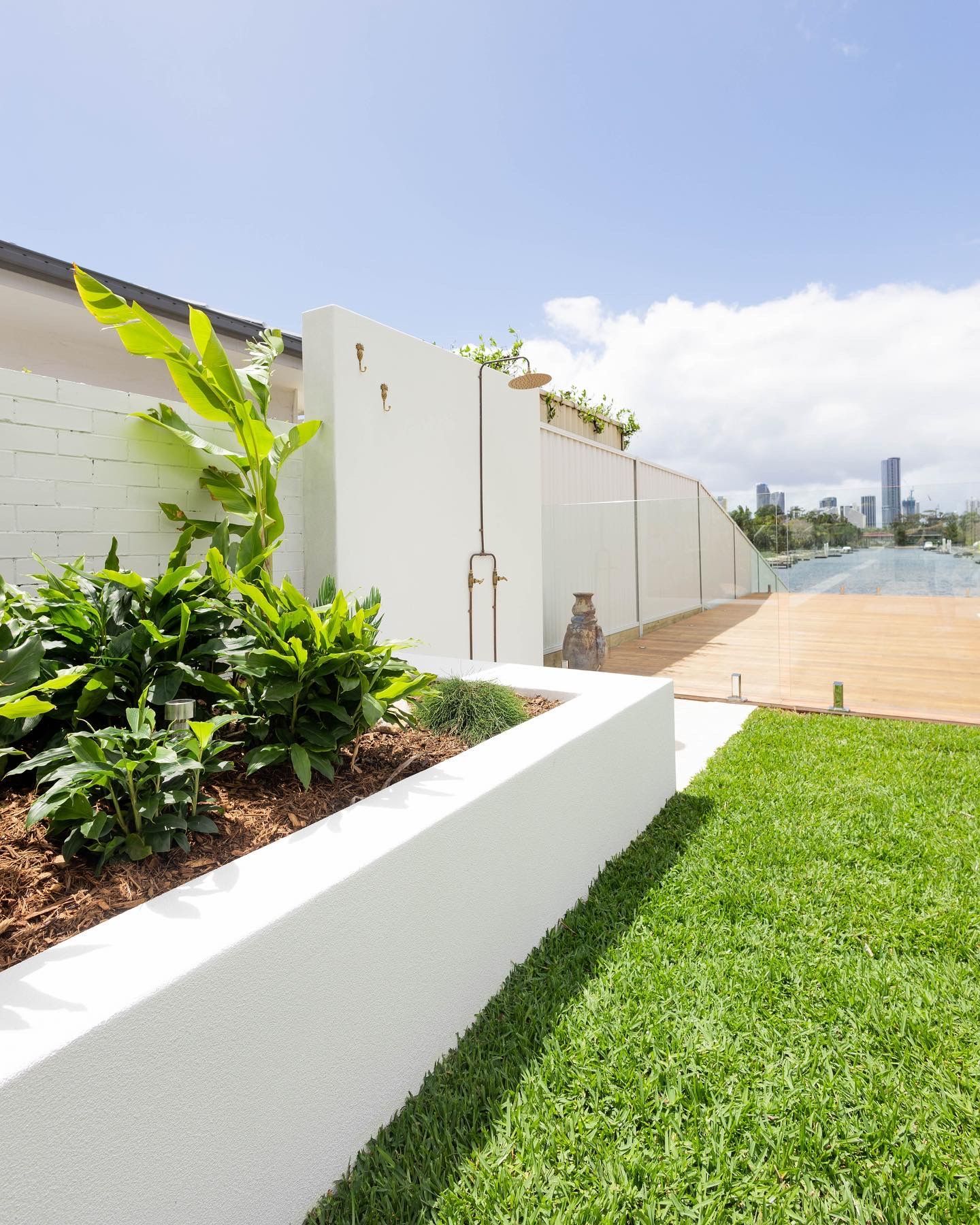 A landscaped garden bed with lush greenery, a white privacy wall with an outdoor shower, and a wooden deck near a canal.