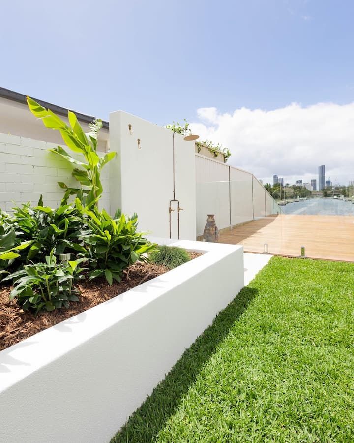 A modern white outdoor shower and garden bed border a lush green lawn, overlooking a river and city skyline.