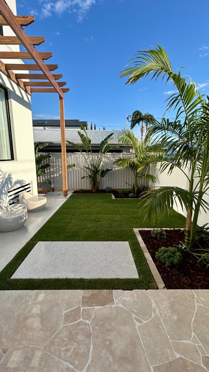 A sunlit backyard with a stone patio, green grass lawn, a square white paver, a wooden pergola, and palm trees.