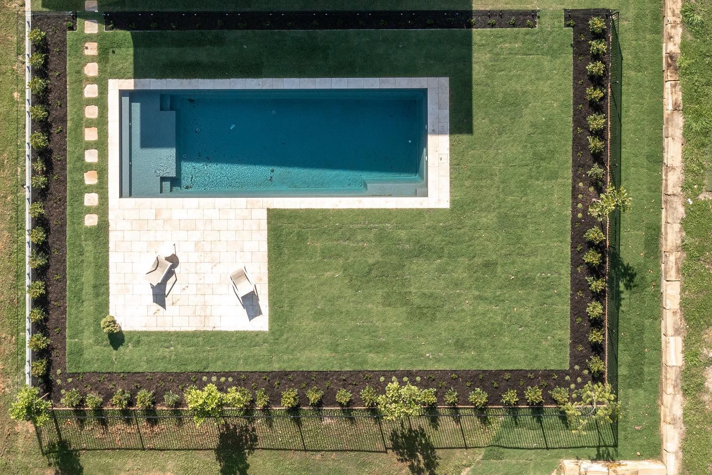 An overhead view of a rectangular swimming pool with a stone patio, surrounded by a lawn and a row of perimeter plants.
