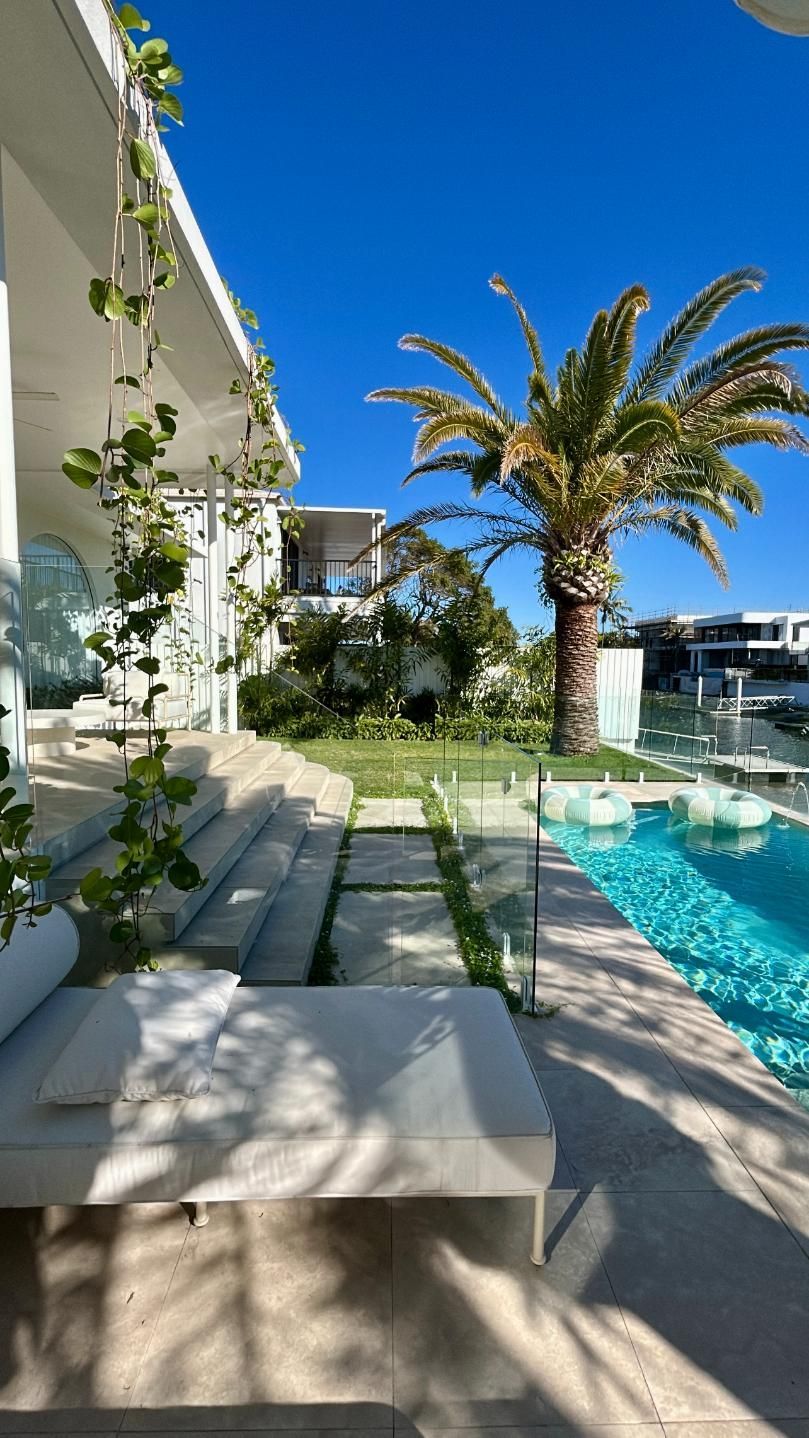 A luxurious patio with a poolside lounge chair, concrete pavers, a lush green lawn, and a palm tree under a blue sky.