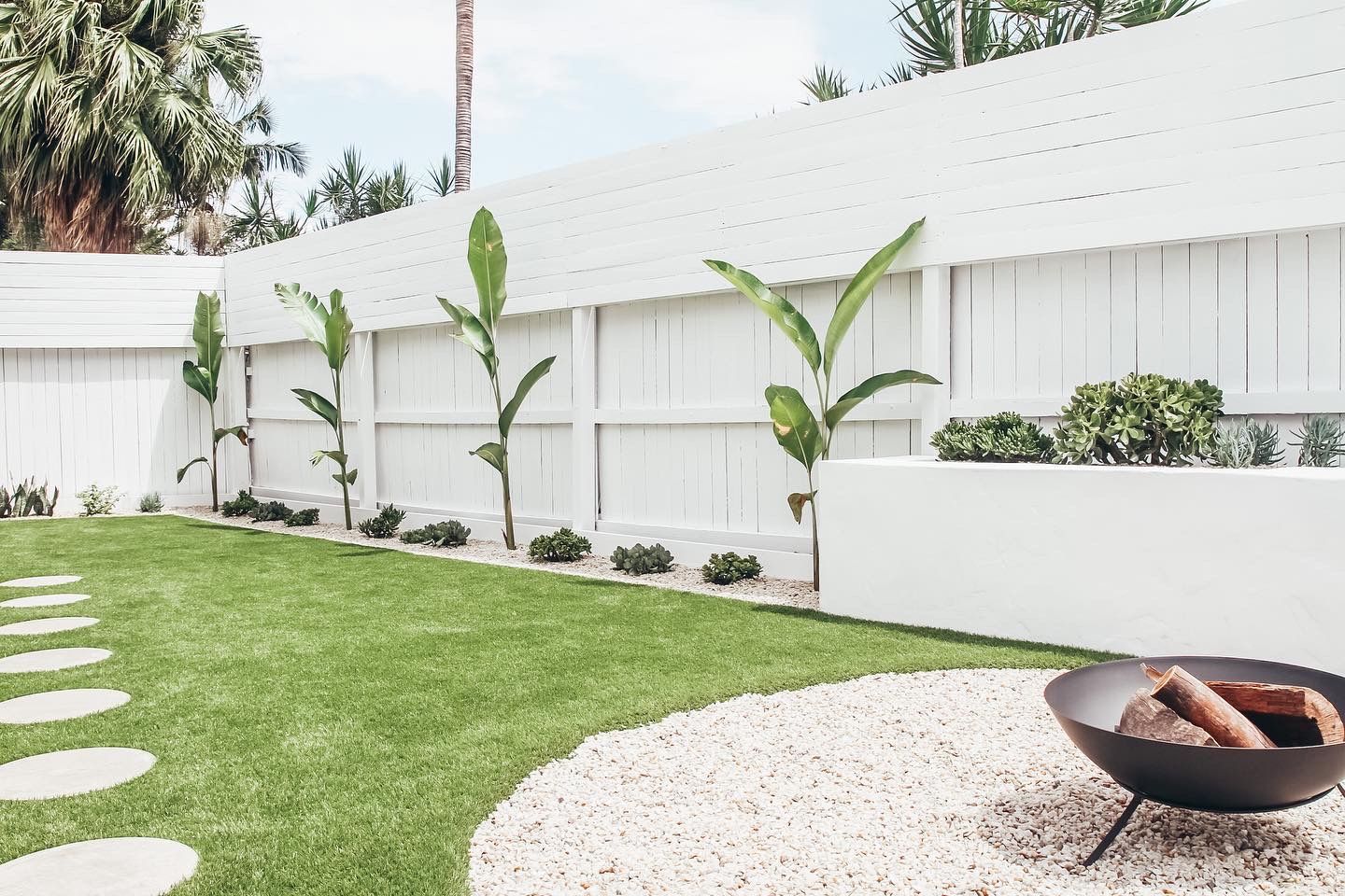 A white backyard fence features young plants, a green lawn with stone path, and a pebble patio with a metal fire pit.