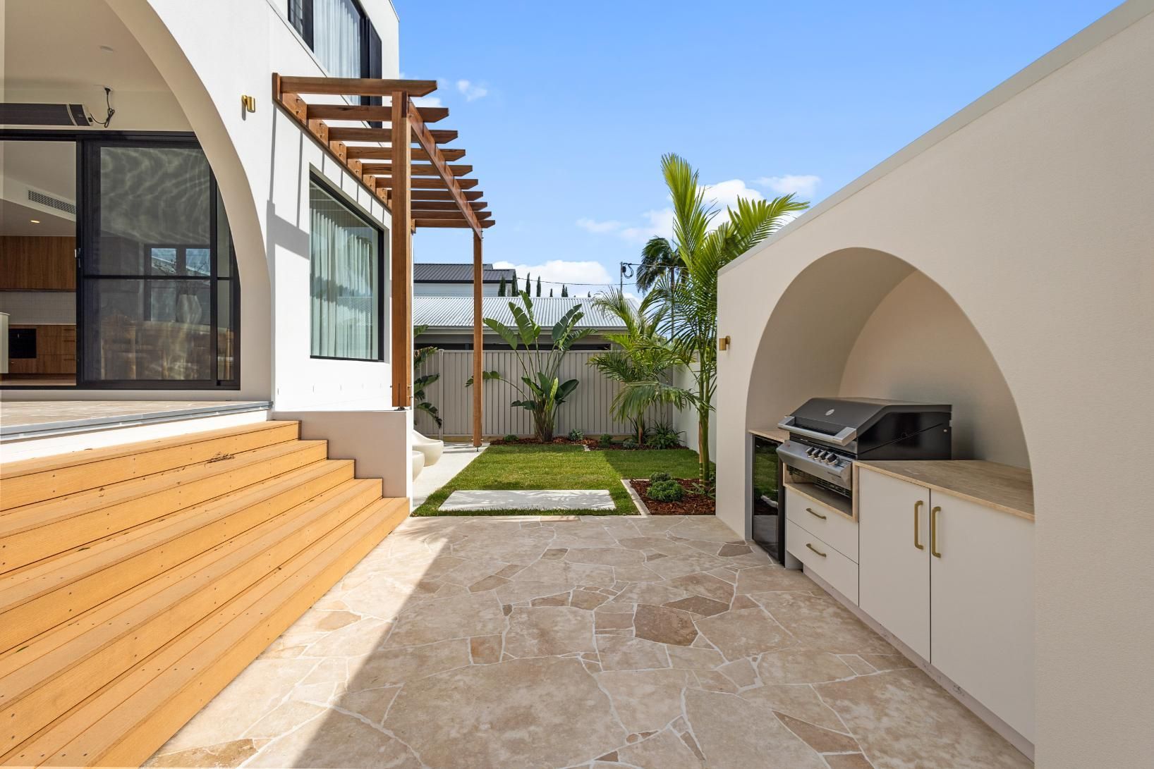 A modern backyard patio features stone flooring, wooden deck steps, an arched outdoor kitchen, and a small grass lawn.