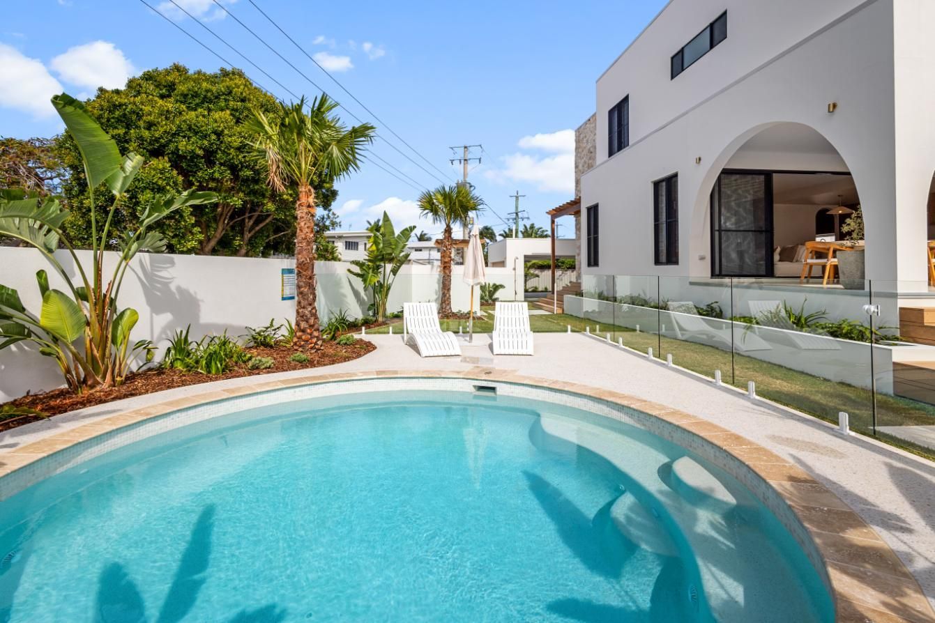 A sunny backyard features a sparkling blue oval pool, two white lounge chairs, and lush greenery next to a white house.