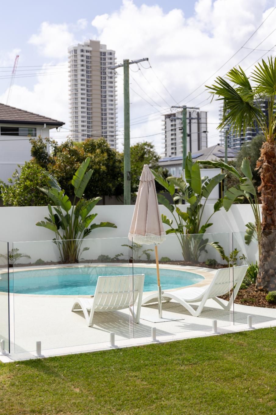 Two white lounge chairs and a closed patio umbrella sit poolside in a backyard with tropical plants and city high-rises.