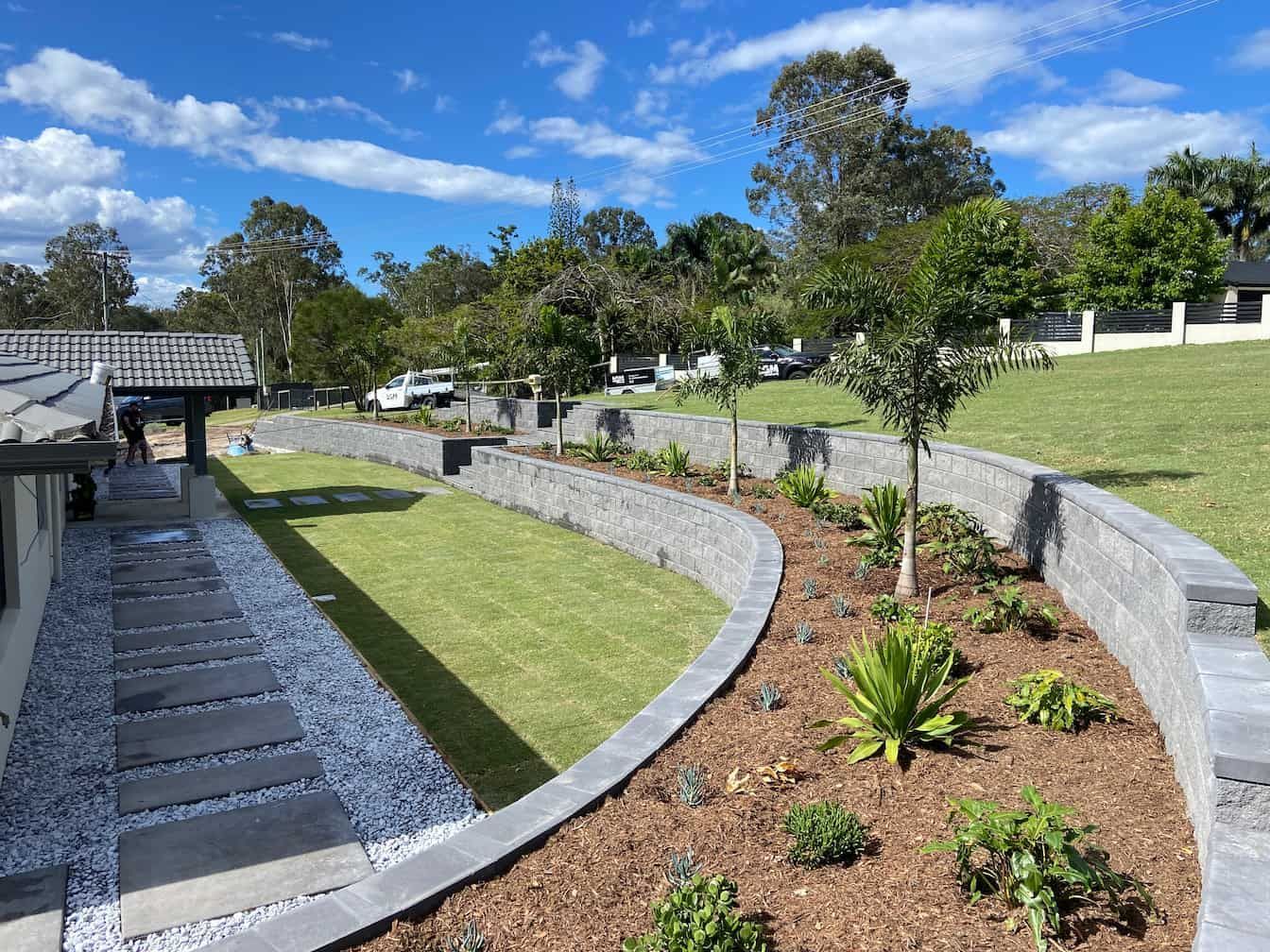 A landscaped yard with a curved stone retaining wall, grass lawn, mulch garden bed, and a stone paver path beside a house.