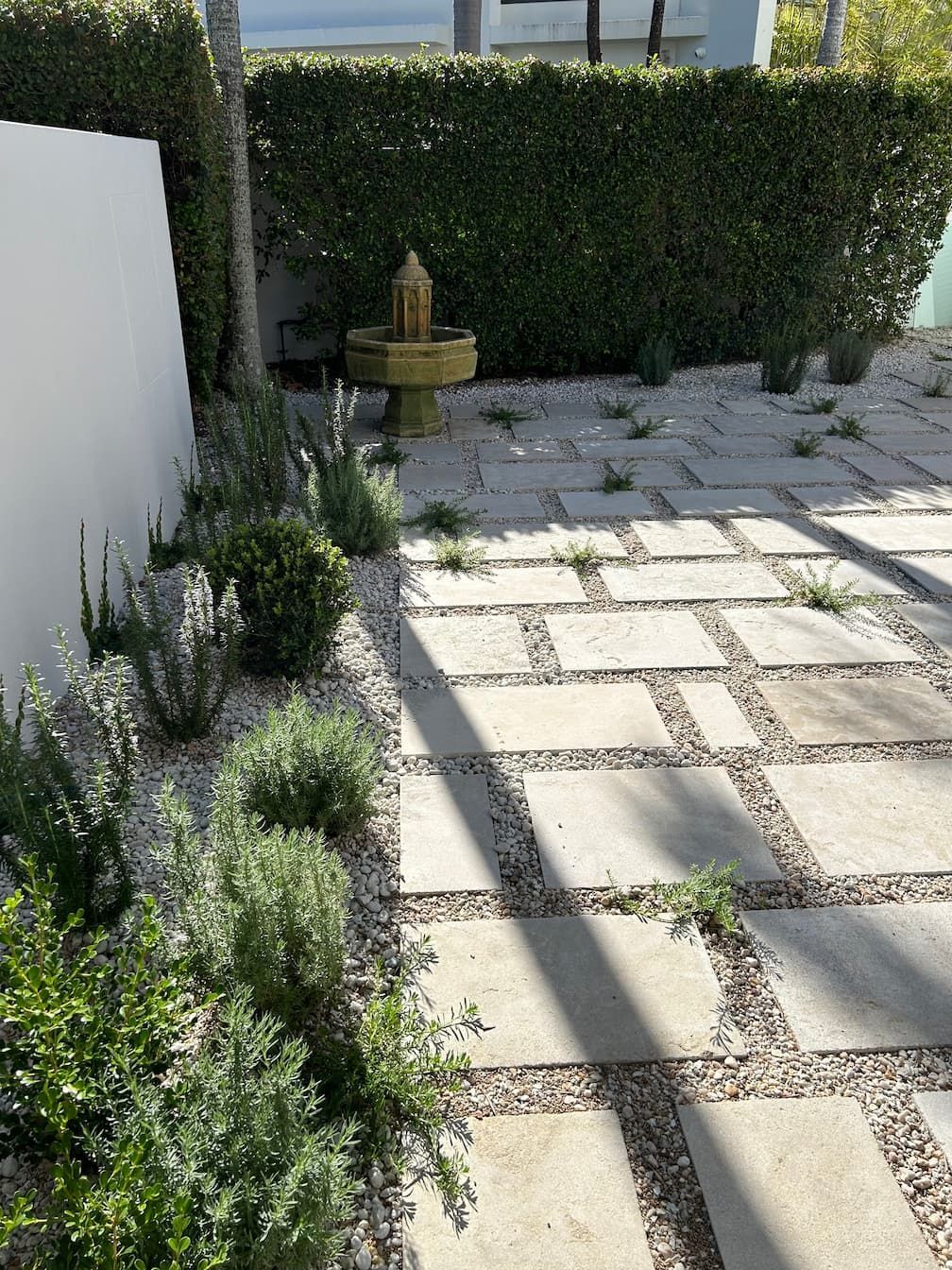 A stone paver walkway with gravel gaps leads to a small garden fountain, flanked by a green hedge and a white wall.