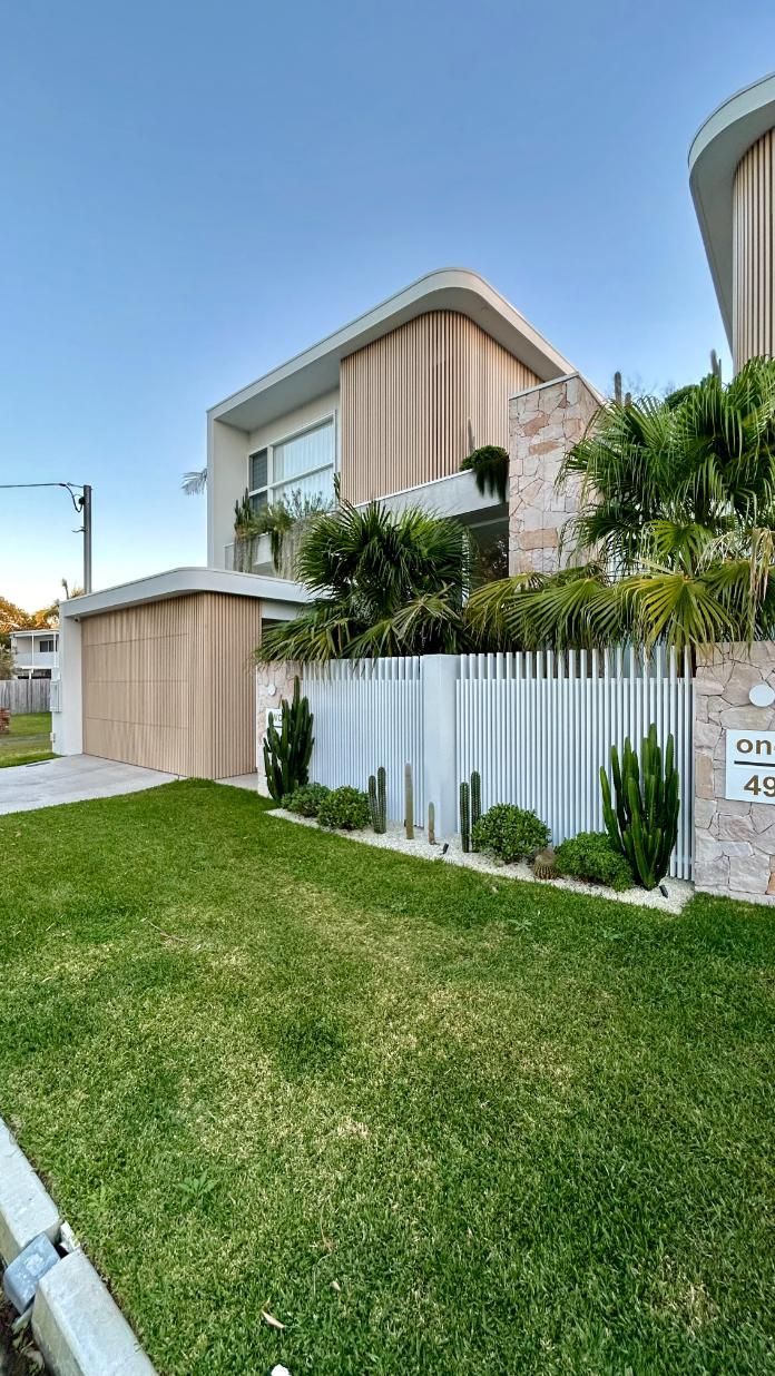 A modern two-story home with light wood panels, a stone pillar, a manicured lawn, and a white vertical-slat fence.