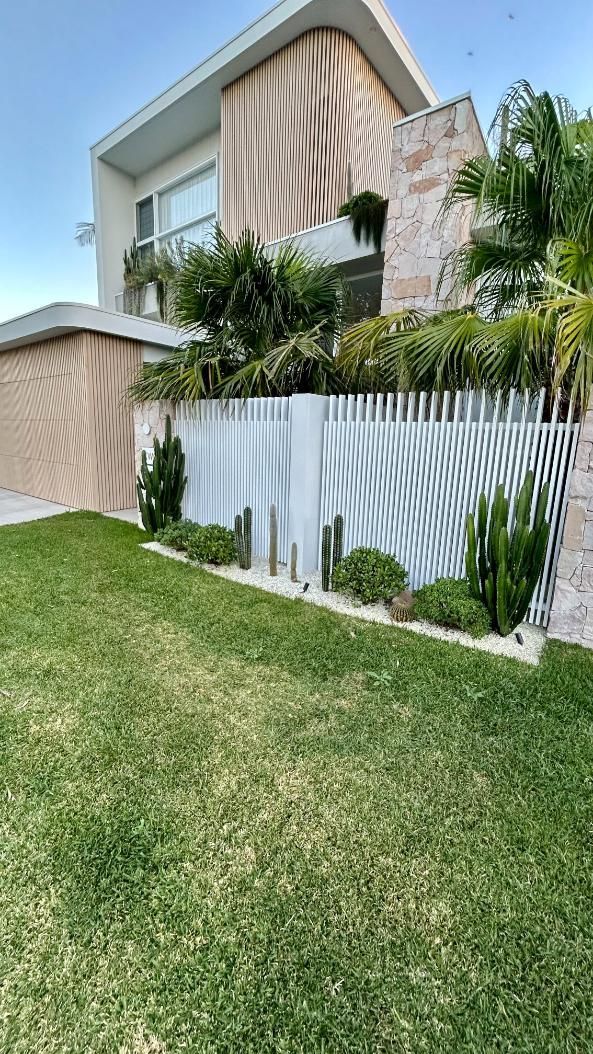 A modern two-story home with a tan textured facade, a white decorative fence, and landscaped greenery in the front yard.