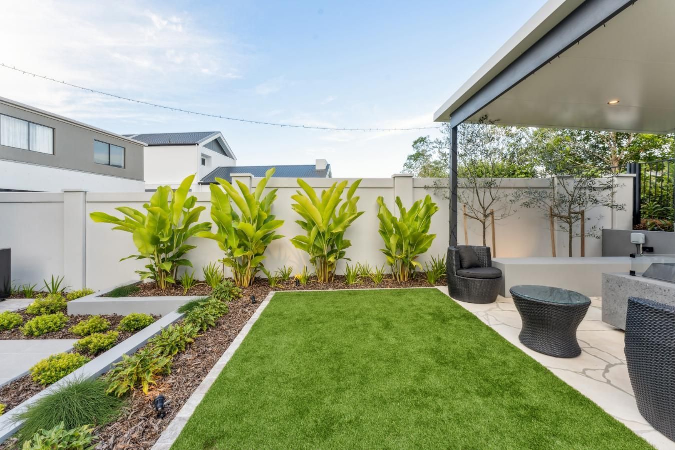 A small, modern backyard with a green lawn, four tall green plants against a light wall, and a covered patio area.