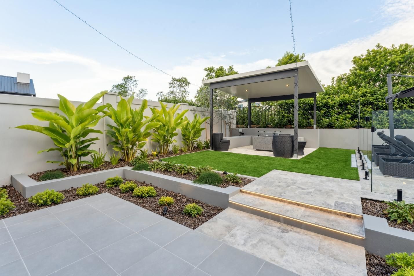 A landscaped backyard featuring a gray-tiled patio, stone steps, a lush lawn, a gazebo, and tiered garden beds with plants.