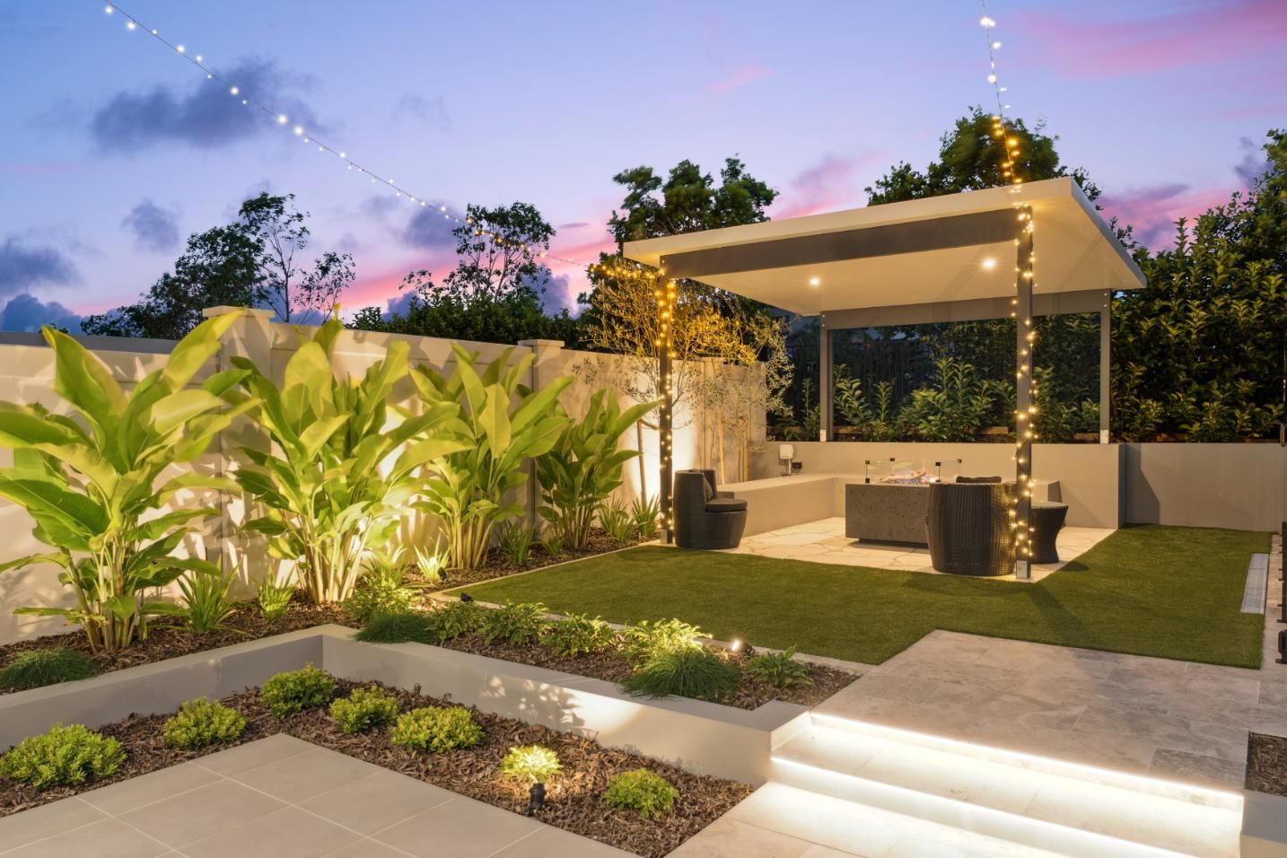 A modern backyard patio at sunset with a lit pergola, green grass, stone steps, and tropical plants.