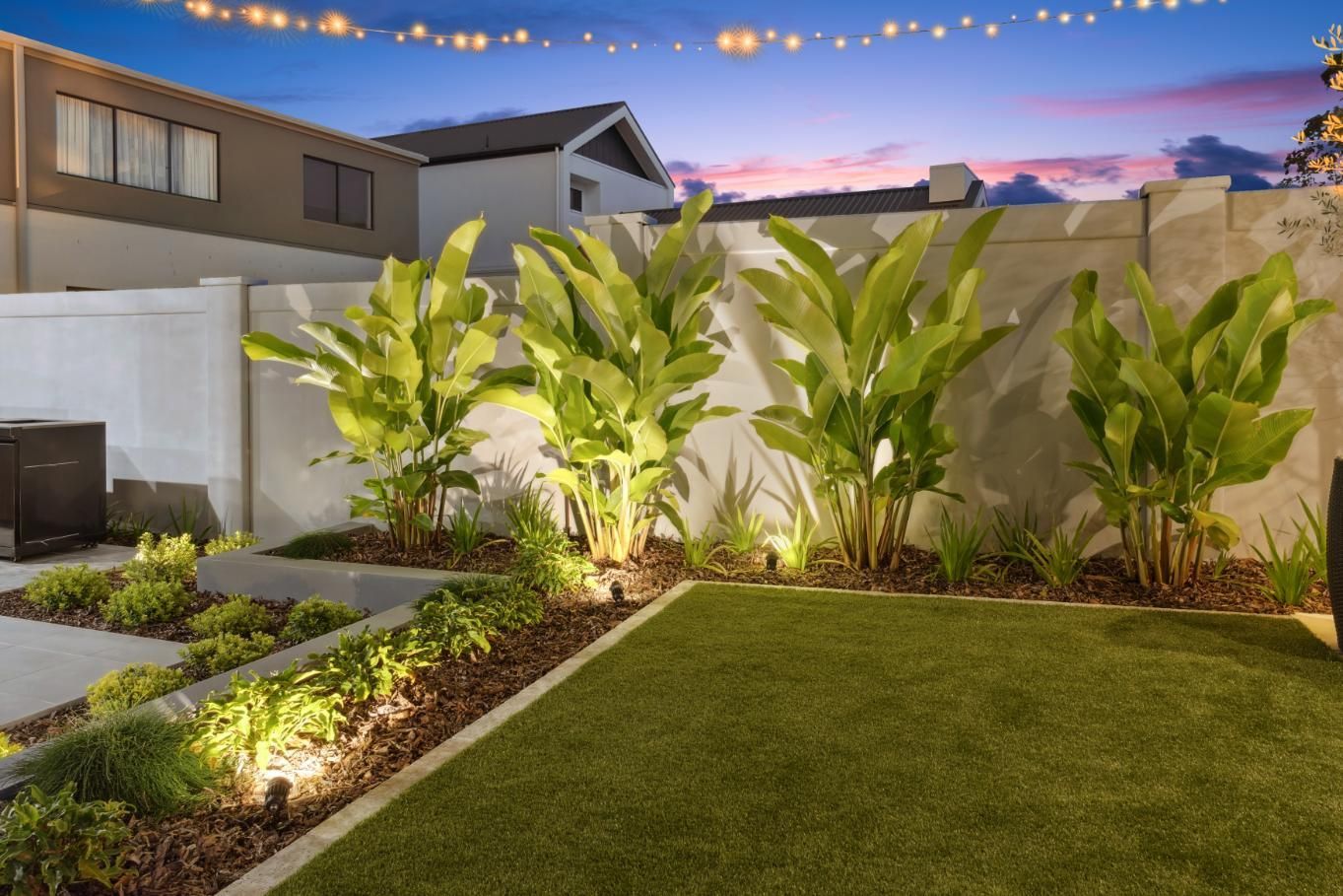 A landscaped backyard at twilight featuring lush green plants, a manicured lawn, and warm decorative string lights.