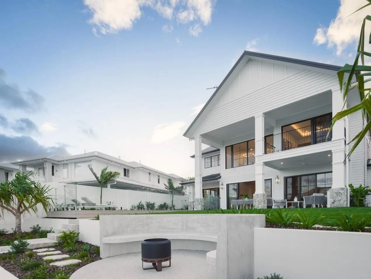 A modern white multi-story house with a backyard featuring a concrete fire pit area, landscaping, and stone pathways.