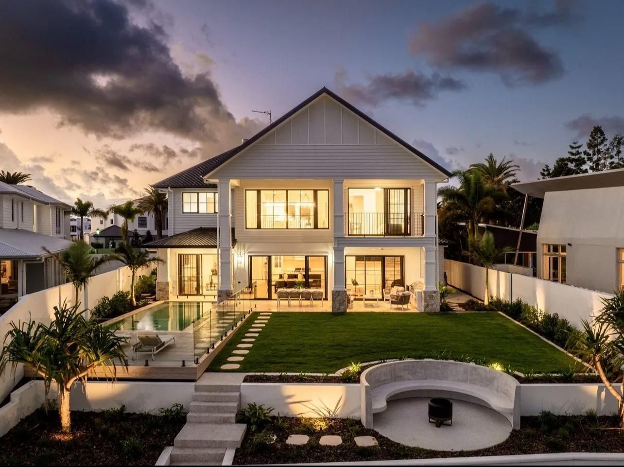 A luxury two-story house at dusk, featuring an illuminated swimming pool, a landscaped backyard, and a curved stone seat.