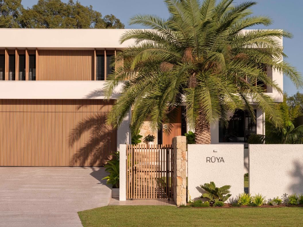 A modern two-story home with light wood-slatted exterior walls, a palm tree, and a stone wall with the word 