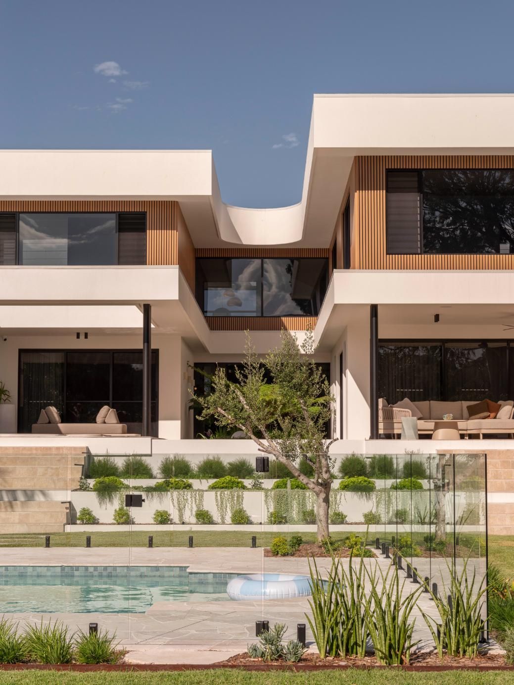 A modern two-story home with a curved white facade, slatted wood accents, tiered landscaping, and a pool in the foreground.