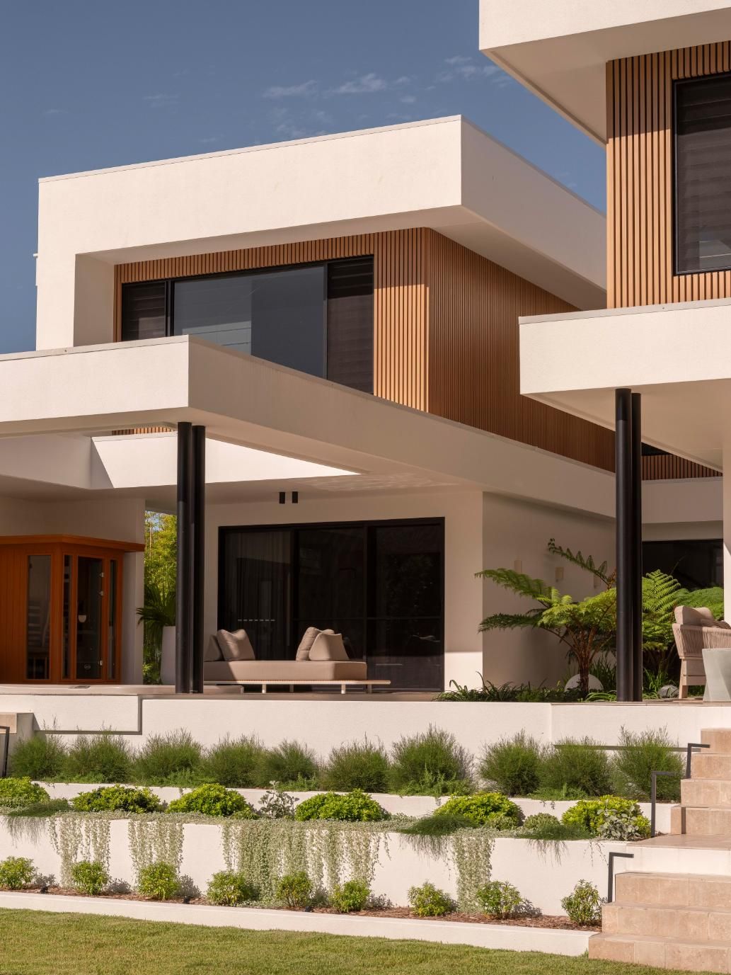 Modern house with white stucco walls, light wood slats, black-framed windows, a patio, and tiered landscaped planters.