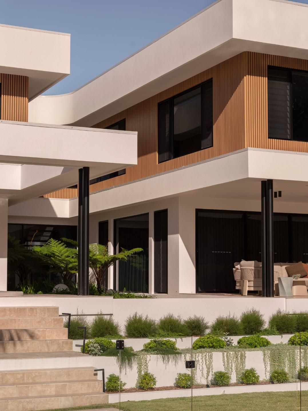 A modern two-story home with white walls, wood paneling, large windows, and a landscaped multi-tiered front patio.