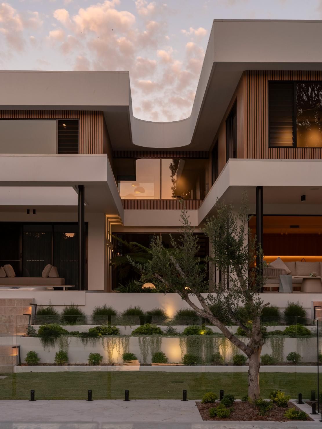 A modern two-story home with clean white architecture and vertical wood slats, set behind a terraced garden at dusk.