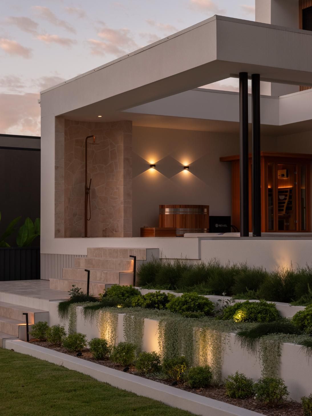 A modern house exterior at twilight, featuring terraced landscaping, illuminated stone walls, and a patio with a shower.
