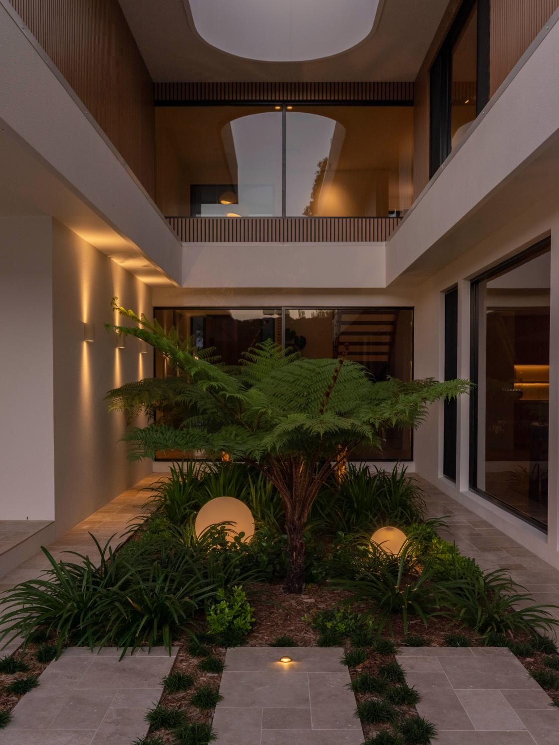 A modern courtyard with a central fern, illuminated by spherical lights at dusk, surrounded by architectural walls.