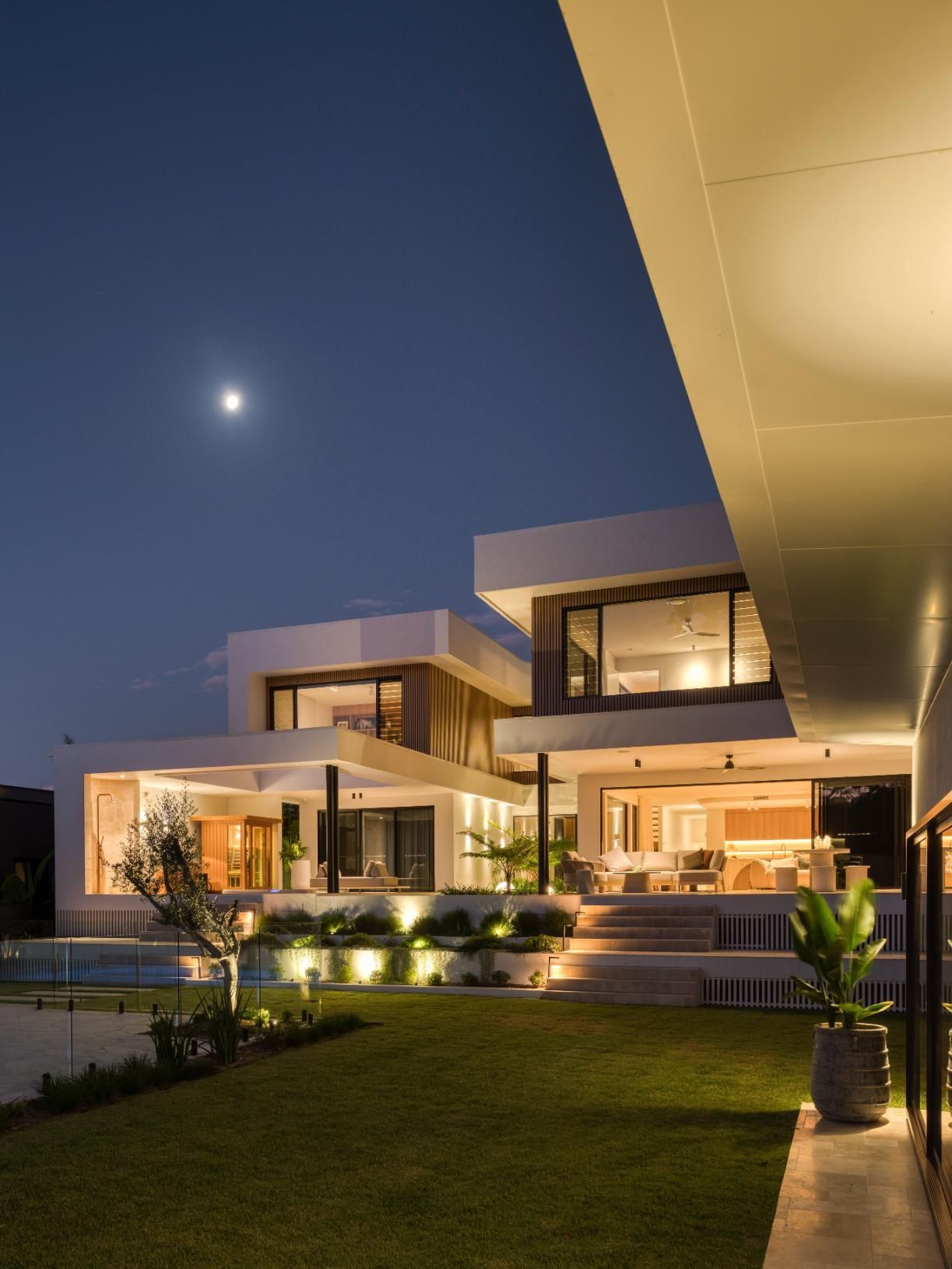 A modern two-story home at night with warm interior lighting, a landscaped lawn, and a clear sky with a visible moon.