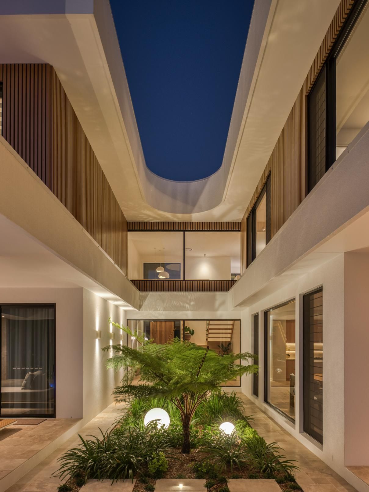 A modern courtyard at night with a central tree, illuminated by ground lights, surrounded by sleek, multi-story architecture.