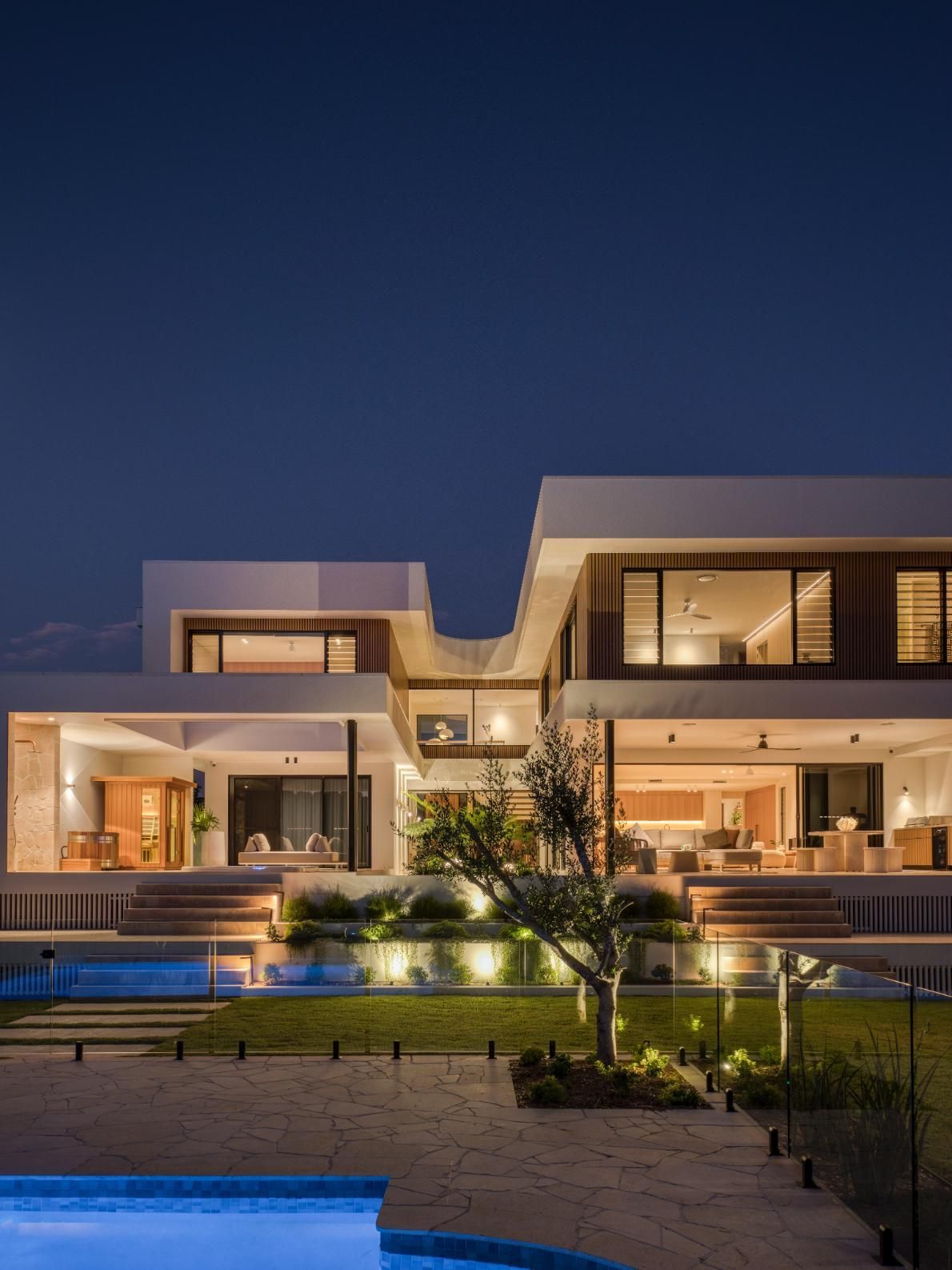 A modern, two-story luxury home at twilight, featuring an illuminated outdoor patio, large windows, and a swimming pool.