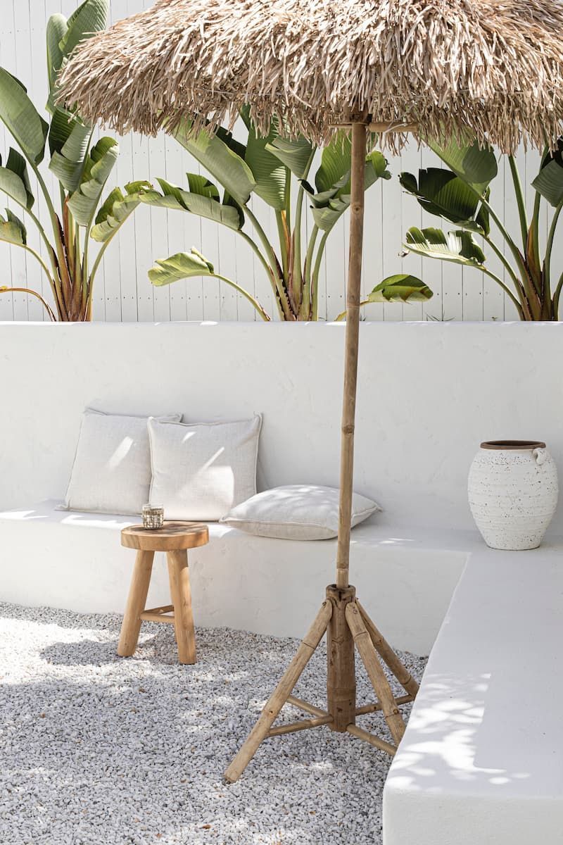 A thatched beach umbrella stands in a gravel patio area next to a white bench with cushions and a small wooden stool.