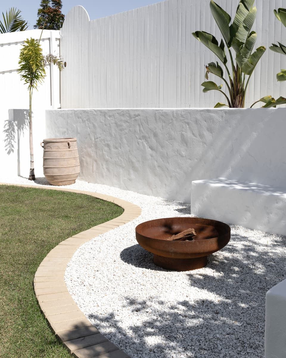 A rustic metal fire pit sits on a gravel patio beside a curved stone wall and lawn in a bright, modern backyard.