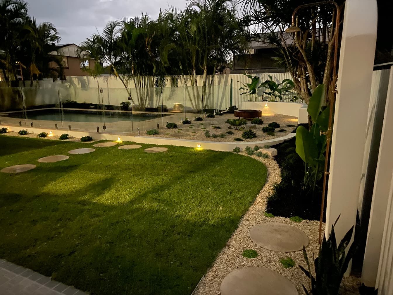 A backyard at twilight featuring a lawn with stone stepping stones, a swimming pool, palm trees, and outdoor lighting.