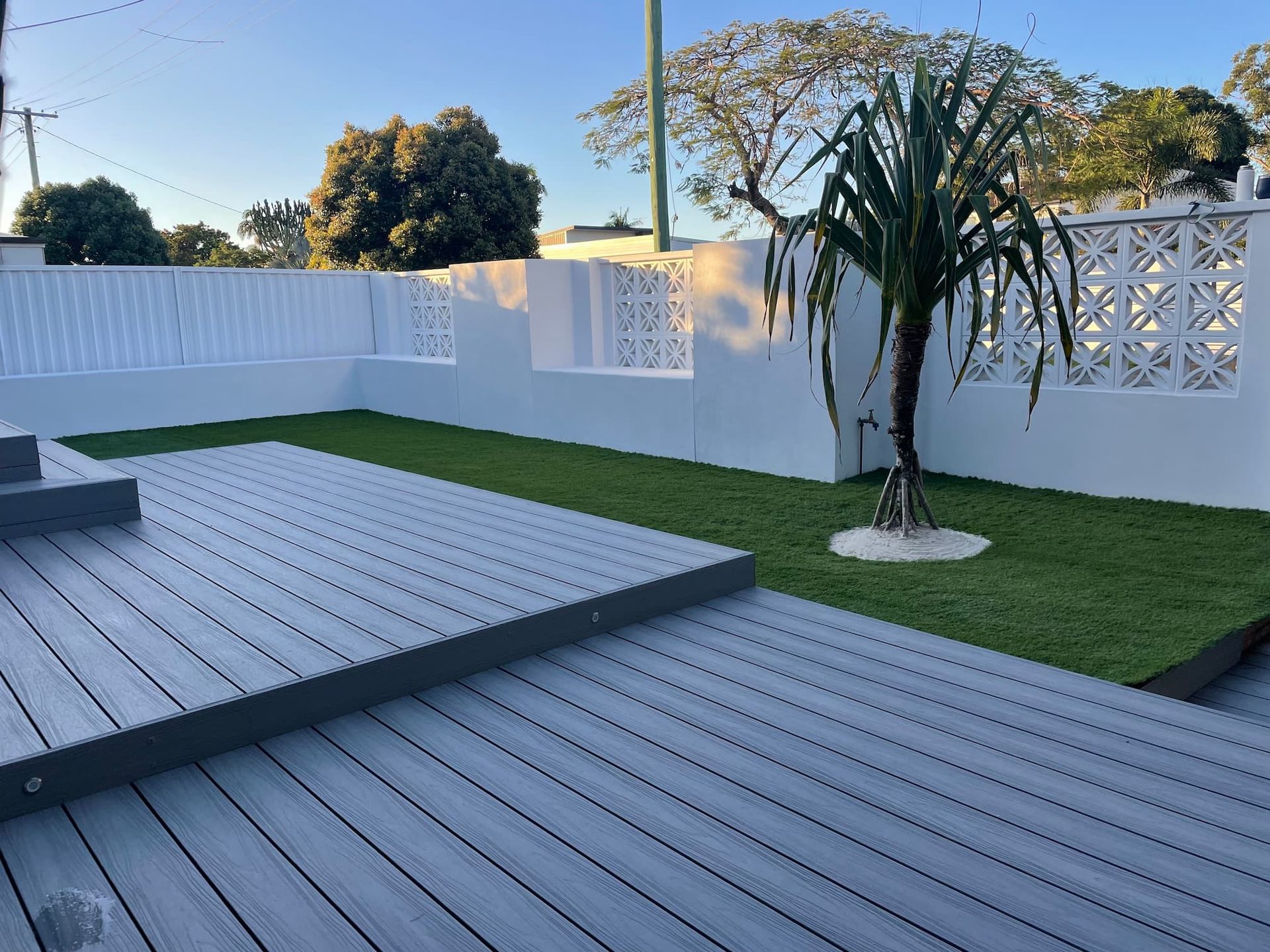 Grey tiered deck leading to a green lawn with a small tree and white decorative wall under a clear blue sky.