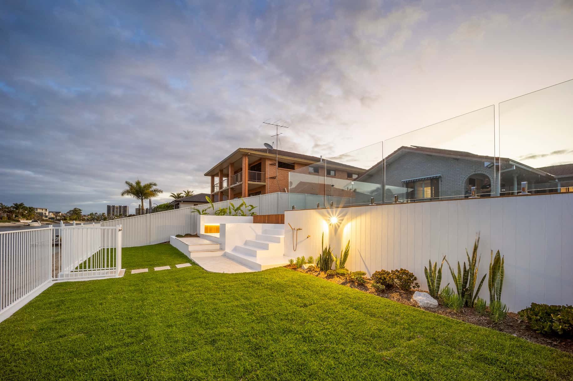 A backyard with green grass, white retaining walls, steps leading to a house, and a glass fence at sunset.